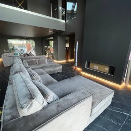 Usługi Elektryczne Dariusz Malenga - Nowoczesne wnętrze z szarym narożnikiem i podświetleniem LED wzdłuż ściany i podłogi z czarnego marmuru. Widoczny karton w niszy ściennej i okno z rusztowaniem na zewnątrz.