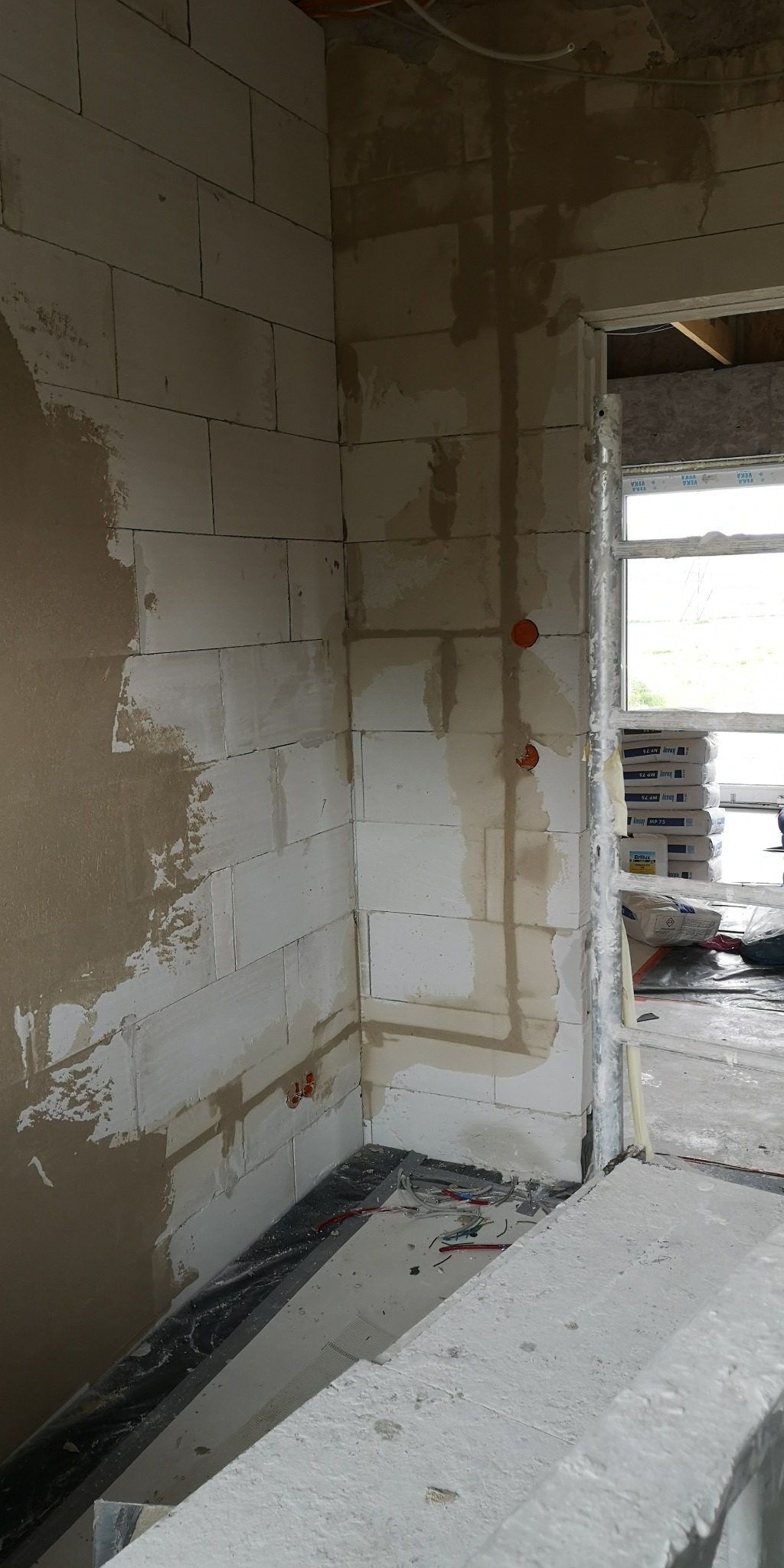 Wnętrze w trakcie prac wykończeniowych: ściany z betonu komórkowego, częściowo otynkowane, z widocznymi puszkami elektrycznymi i przewodami. Okno z workami zaprawy w tle.