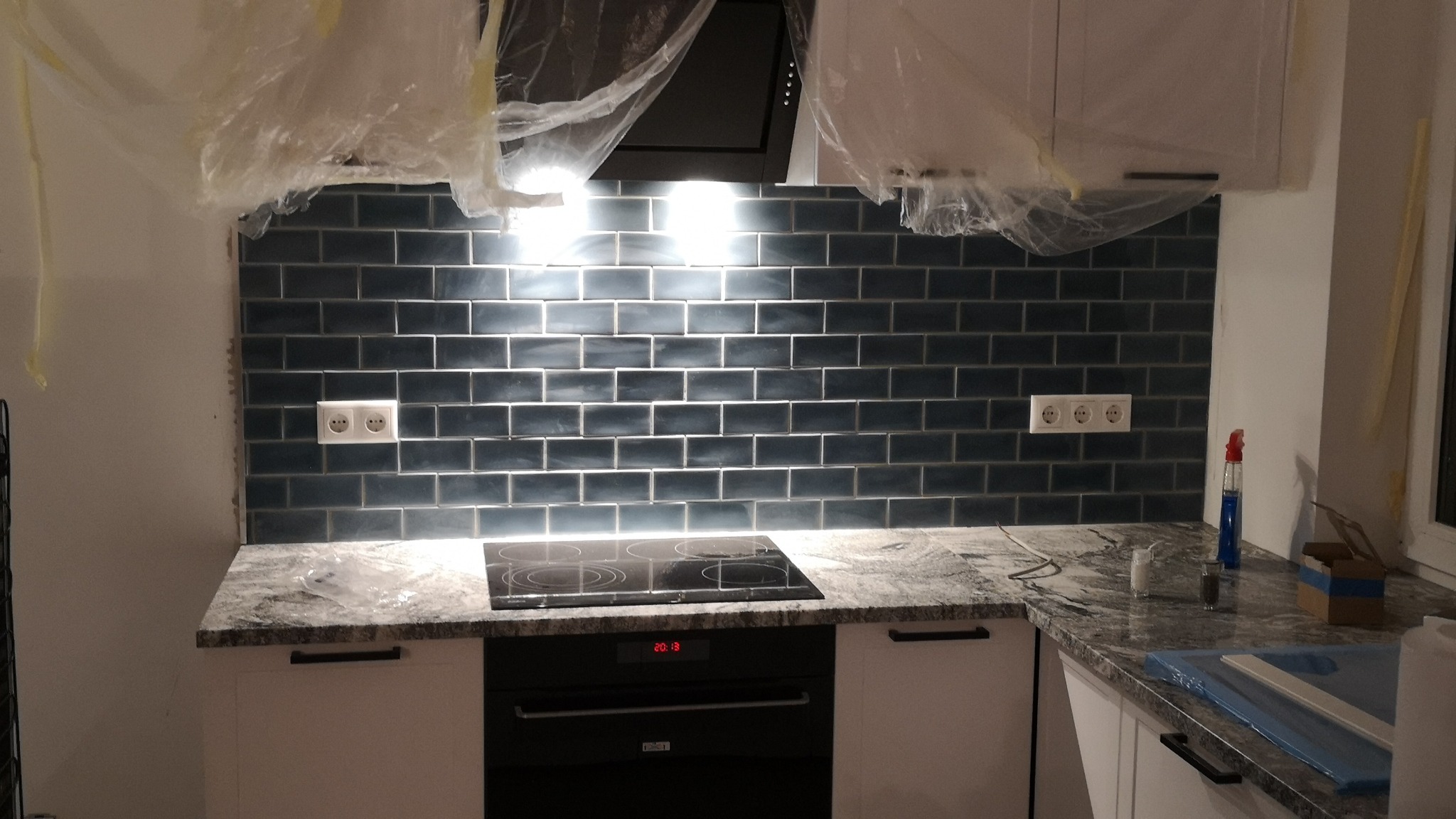 Błyszczące, ciemne płytki ułożone na ścianie nad blatem kuchennym z zamontowanymi gniazdkami elektrycznymi. Widoczna płyta indukcyjna i okap. Częściowo zakryte folią ochronną.