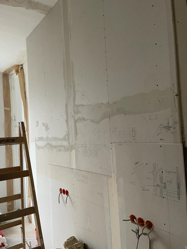 Ściana z płyt gipsowo-kartonowych w trakcie budowy, z widocznymi szkicami instalacji elektrycznej i gniazdkami. Ustawiona drabina. Surowy stan wykończenia wnętrza.