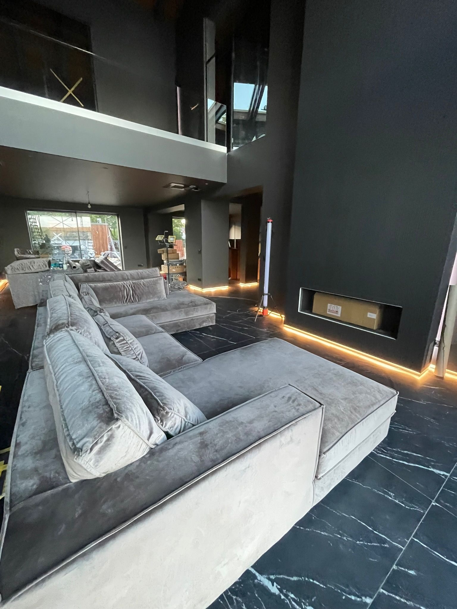 Nowoczesne wnętrze z szarym narożnikiem i podświetleniem LED wzdłuż ściany i podłogi z czarnego marmuru. Widoczny karton w niszy ściennej i okno z rusztowaniem na zewnątrz.