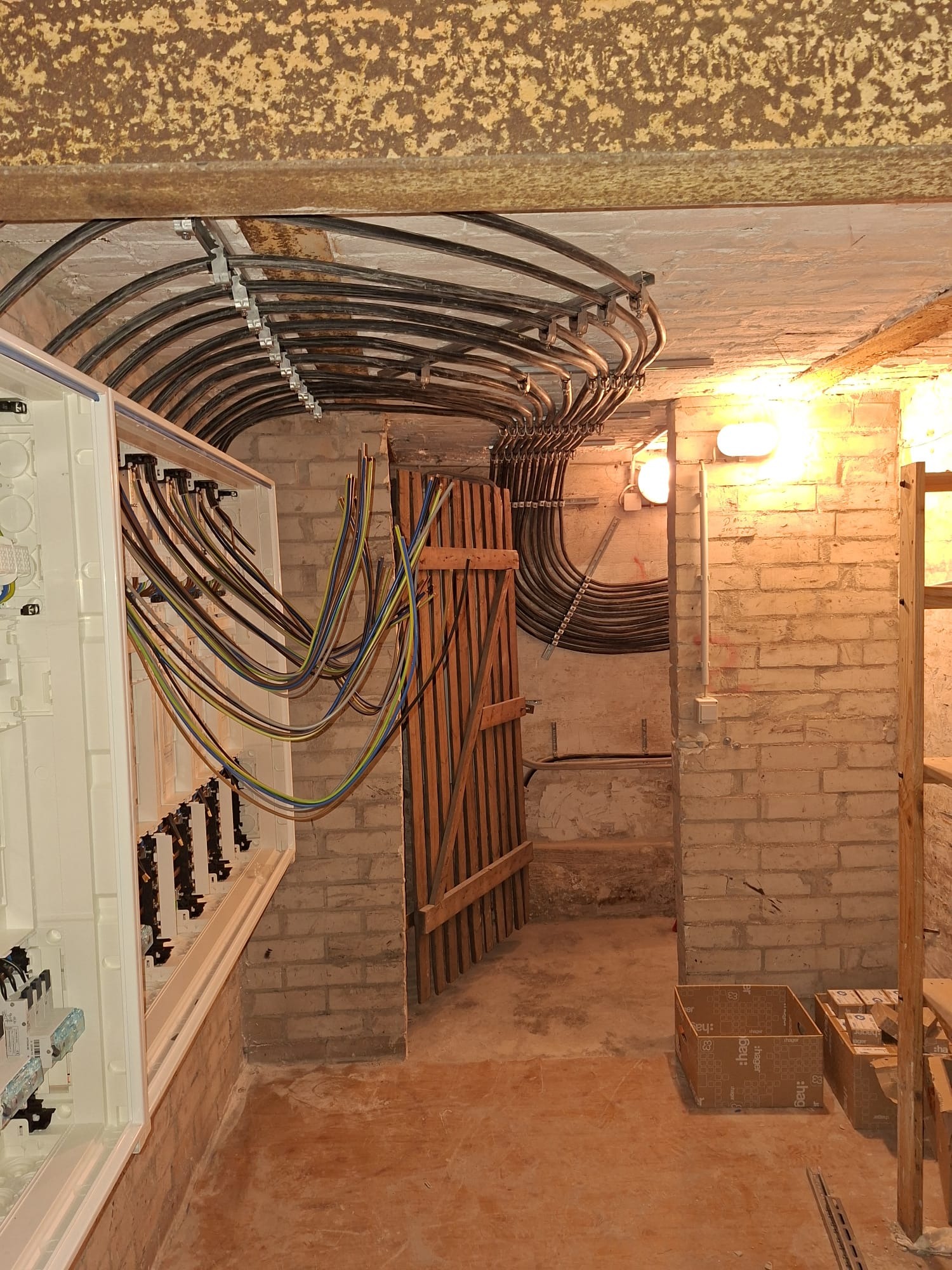 Rozdzielnia elektryczna w kamienicy z widocznymi kablami ułożonymi pod sufitem i skrzynką rozdzielczą na ścianie z cegły. Widoczne kable w kolorowej izolacji.