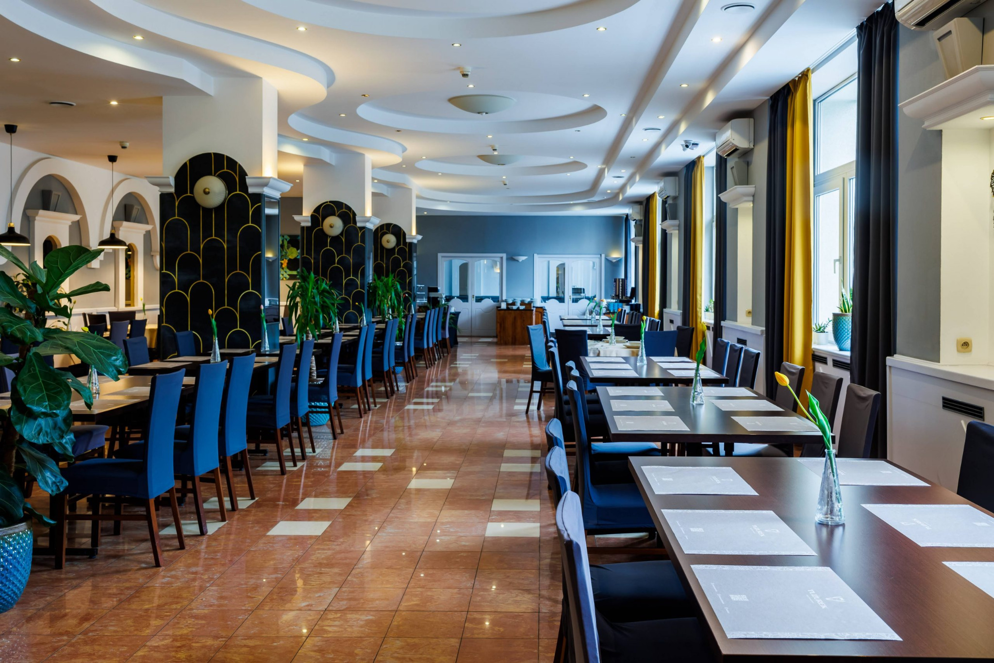 Elegancka sala restauracyjna z długimi stołami nakrytymi dla gości, krzesła w odcieniach granatu, dekoracyjne kolumny i rośliny doniczkowe. Przygotowana przestrzeń na event.