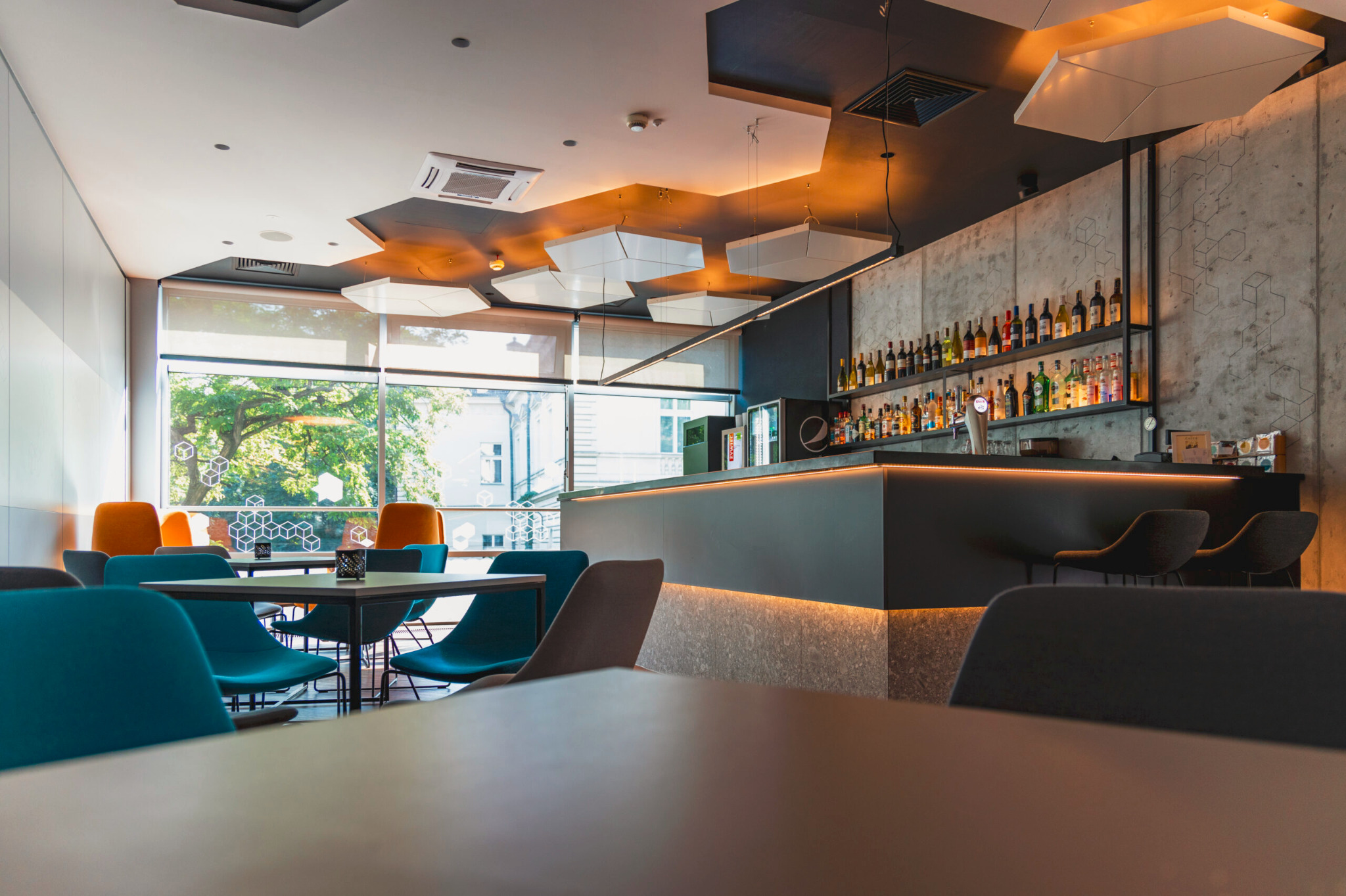 Nowoczesne wnętrze baru z oświetlonym barem, rzędem butelek na półce i stolikami z kolorowymi krzesłami. Duże okno z widokiem na zieleń. Geometryczne lampy sufitowe.