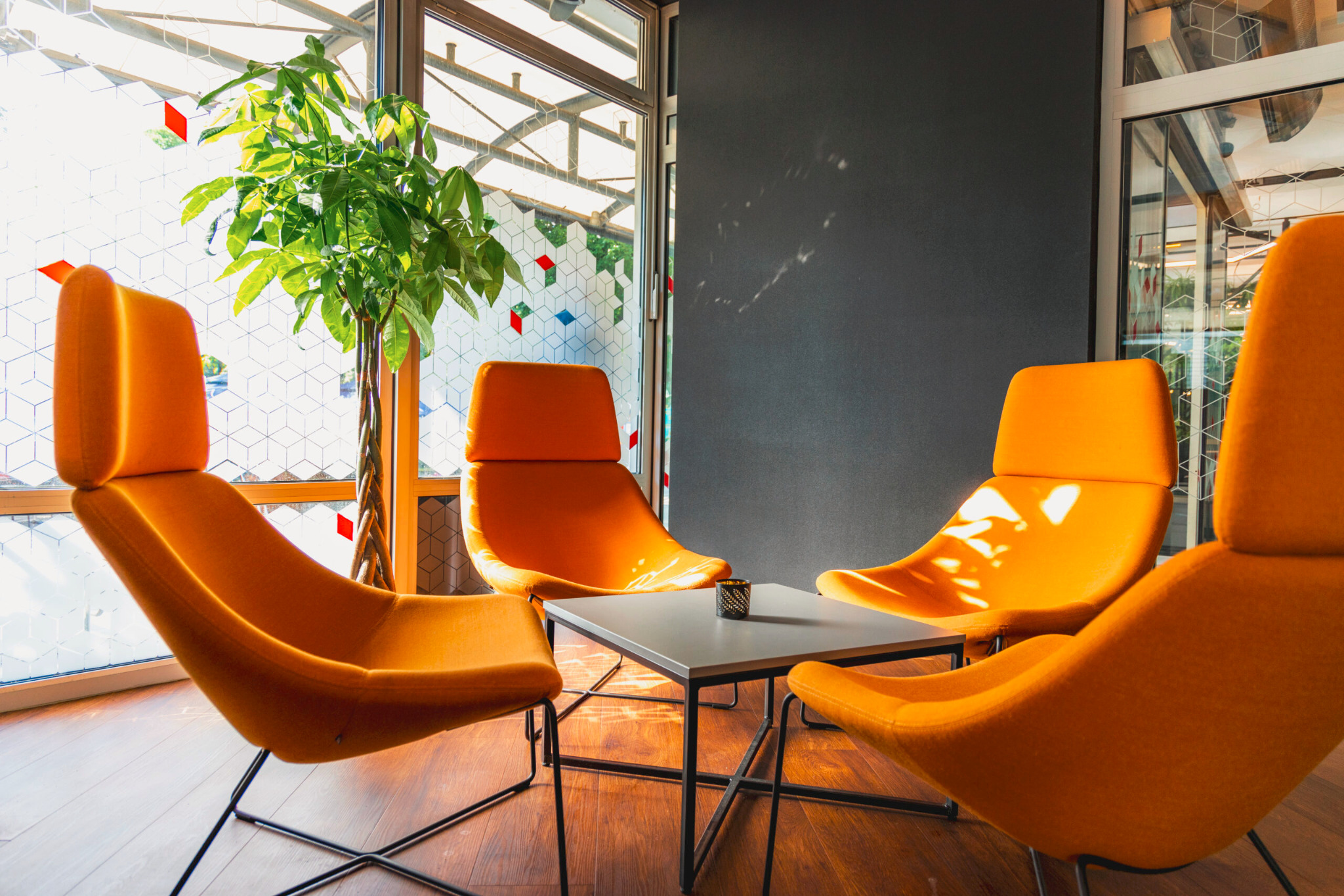 Nowoczesna aranżacja wnętrza z czterema pomarańczowymi fotelami i stolikiem kawowym, doświetlona naturalnym światłem wpadającym przez okno z geometrycznym wzorem.