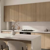 Minimalistyczna kuchnia z drewnianymi szafkami, białym blatem i wyspą. Wazon z suchymi gałązkami dodaje naturalnego akcentu. Nowoczesny design, jasne wnętrze.