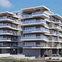 Nowoczesny apartamentowiec z balkonami i szklanymi balustradami, widok z dołu, błękitne niebo w tle, zieleń u podnóża budynku, auta na parkingu.