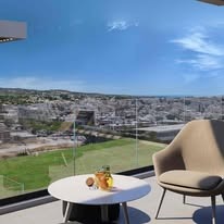 Nowoczesny balkon z widokiem na miasto: beżowe krzesło, okrągły stolik z napojami, szklana balustrada. Słoneczny dzień, błękitne niebo, panorama miejska w tle.