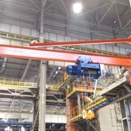 Suwnica konsolowa z wciągnikiem linowym ELMOT w hali przemysłowej. Pomarańczowa konstrukcja przymocowana do betonowego filaru, niebieski wciągnik. Widoczne elementy konstrukcji hali.