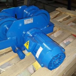 Niebieski wciągnik linowy ELMOT z silnikiem, na drewnianej palecie. Widoczne przekładnie zębate i tabliczka znamionowa. Urządzenie do transportu bliskiego w zakładach.