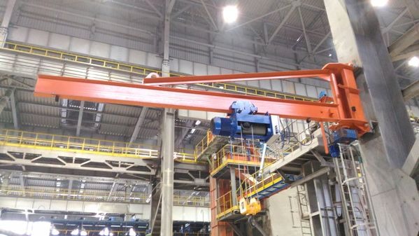 Suwnica konsolowa z wciągnikiem linowym ELMOT w hali przemysłowej. Pomarańczowa konstrukcja przymocowana do betonowego filaru, niebieski wciągnik. Widoczne elementy konstrukcji hali.