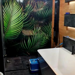 JG BALTIC SPÓŁKA Z OGRANICZONĄ ODPOWIEDZIALNOŚCIĄ - Nowoczesna łazienka z wanną, czarną armaturą i ścianą z motywem tropikalnych liści. Widoczna podłoga z ciemnych płytek i oświetlenie punktowe.