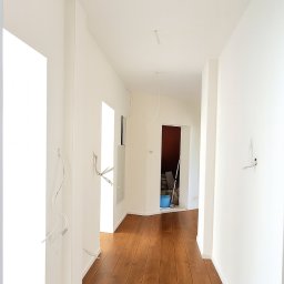 JG BALTIC SPÓŁKA Z OGRANICZONĄ ODPOWIEDZIALNOŚCIĄ - Korytarz po remoncie z drewnianą podłogą i białymi ścianami. Widoczne wystające kable elektryczne, sugerujące prace wykończeniowe w toku. Perspektywa z głębi pomieszczenia.