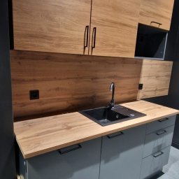 JG BALTIC SPÓŁKA Z OGRANICZONĄ ODPOWIEDZIALNOŚCIĄ - Nowoczesna kuchnia z szarymi szafkami i drewnianymi blatami. Czarny zlew i bateria. Górne szafki z drewnianym fornirem. Minimalistyczny design, czarne uchwyty.