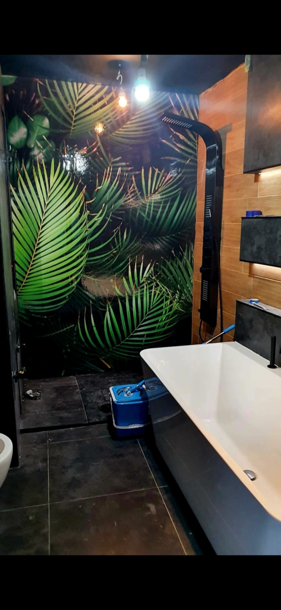 Nowoczesna łazienka z wanną, czarną armaturą i ścianą z motywem tropikalnych liści. Widoczna podłoga z ciemnych płytek i oświetlenie punktowe.