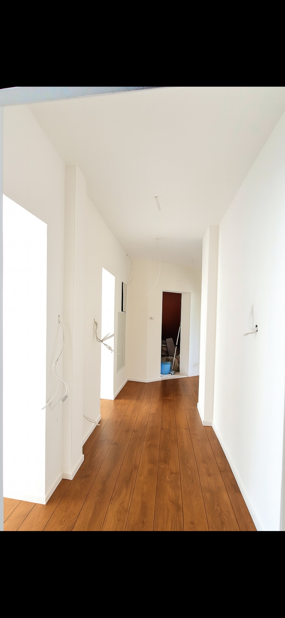 Korytarz po remoncie z drewnianą podłogą i białymi ścianami. Widoczne wystające kable elektryczne, sugerujące prace wykończeniowe w toku. Perspektywa z głębi pomieszczenia.