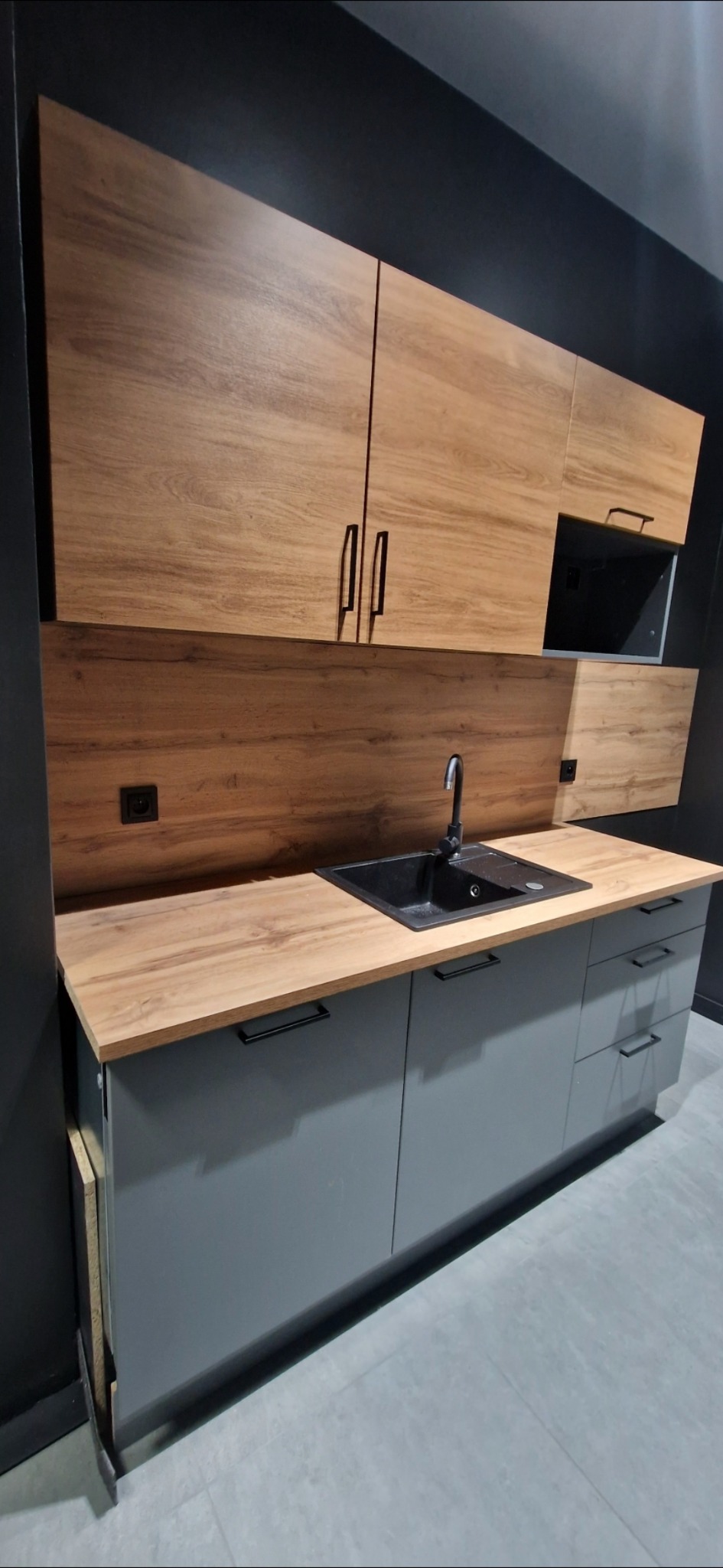 Nowoczesna kuchnia z szarymi szafkami i drewnianymi blatami. Czarny zlew i bateria. Górne szafki z drewnianym fornirem. Minimalistyczny design, czarne uchwyty.