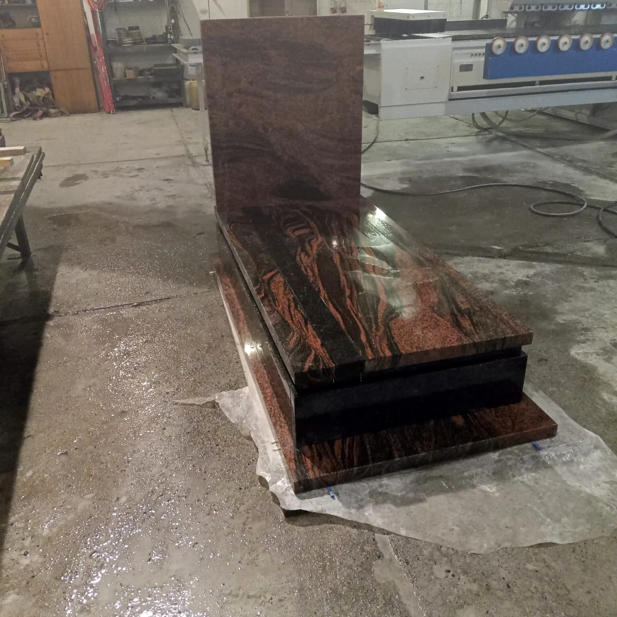 Nowy, dwuczęściowy nagrobek z czerwono-czarnego granitu na folii ochronnej w warsztacie kamieniarskim. Widoczna maszyna do obróbki kamienia.