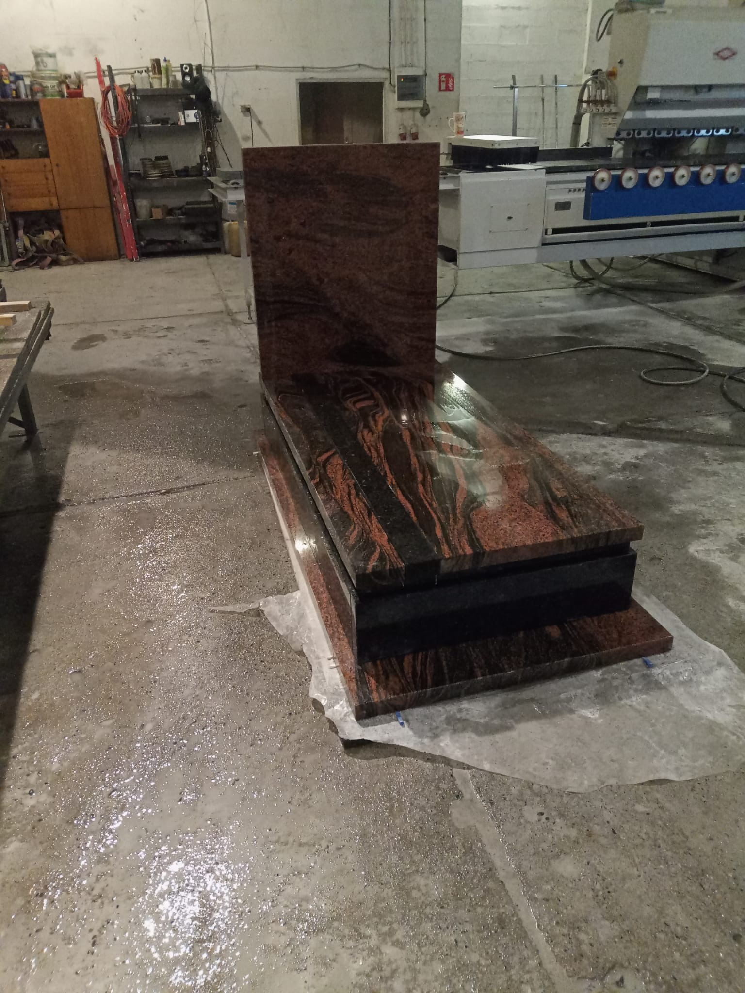 Elementy z granitu w odcieniach czerwieni i czerni, prawdopodobnie części składowe pomnika, leżące na folii w warsztacie kamieniarskim. W tle maszyna do obróbki kamienia.