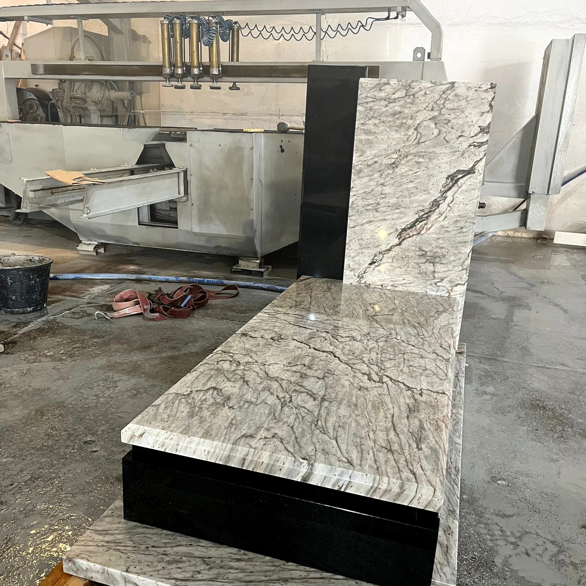 Elementy nagrobka z jasnego i czarnego kamienia, obrobione na maszynie kamieniarskiej. Widoczne pasy transportowe i wiadro. Szlifowany blat z unikalnym wzorem.