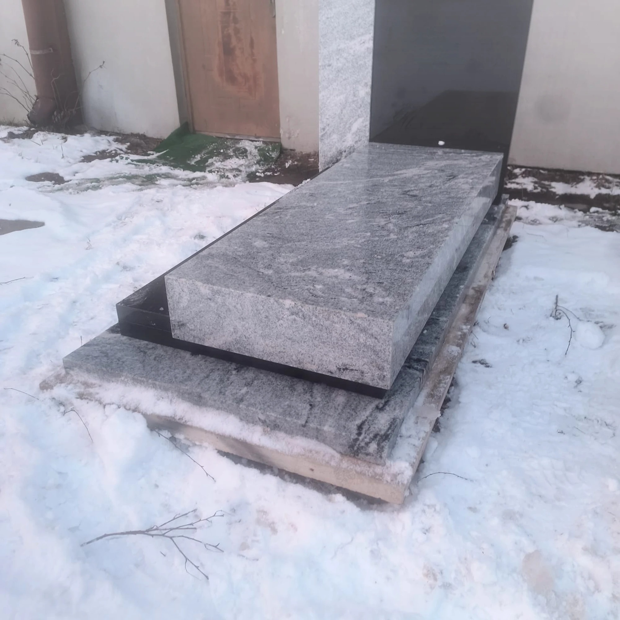 Szary, granitowy element pomnika w zimowej scenerii, z widoczną strukturą kamienia i czarnym wykończeniem, leżący na drewnianych paletach w śnieżnym otoczeniu.