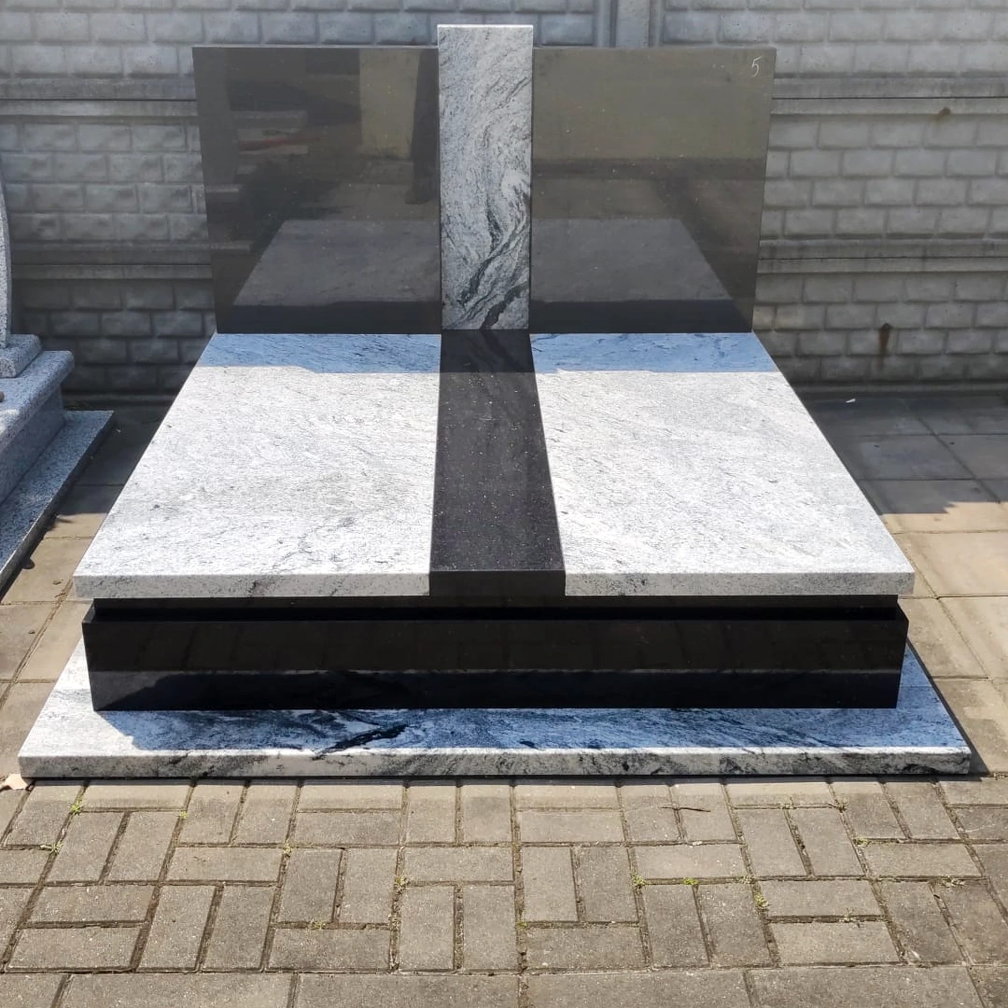 Nowoczesny pomnik z jasnego granitu z czarnymi elementami w kształcie krzyża, na tle szarego muru. Widoczne detale wykończenia i faktura kamienia.