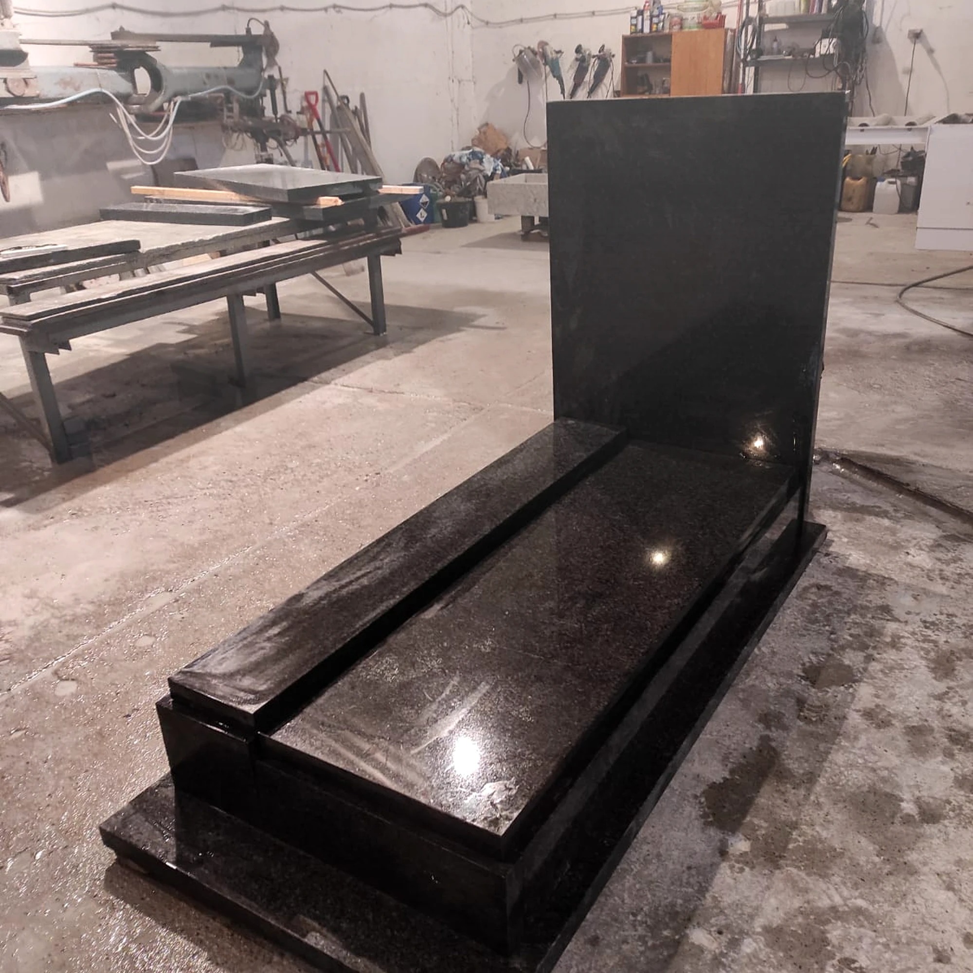 Czarny, błyszczący nagrobek z granitu w warsztacie kamieniarskim. Widoczne narzędzia i elementy obróbki kamienia w tle. Surowy, industrialny charakter.