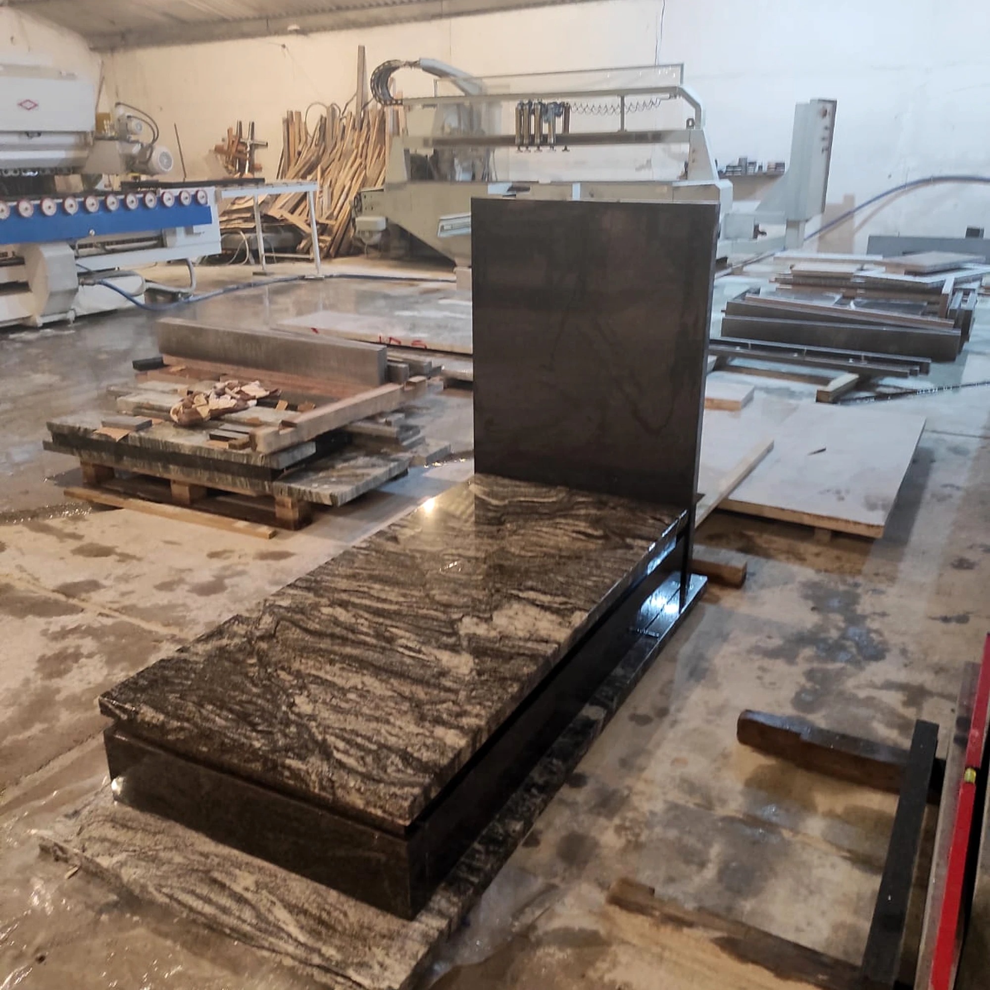 Częściowo złożony, polerowany pomnik z ciemnego granitu w warsztacie kamieniarskim. W tle maszyny do obróbki kamienia i elementy nagrobków.