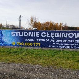 Baner reklamowy z napisem 'Studnie głębinowe' i numerem telefonu na betonowym ogrodzeniu, otoczony zielenią i drzewami w tle.