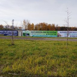 Banery reklamowe: studnie głębinowe, oczyszczalnie ścieków i pralnia dywanów. Widok na łąkę i drzewa w tle. Tablice z numerami telefonów.