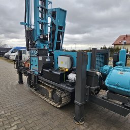 BioTech Aneta Telążka - Mobilna wiertnica studni głębinowych na gąsienicach, w kolorze niebiesko-czarnym, zaparkowana na kostce brukowej. Widoczne elementy sterujące i przewody hydrauliczne.