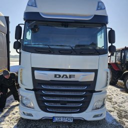 BioTech Aneta Telążka - Biała ciężarówka DAF z przodu, z widoczną tablicą rejestracyjną i osobą sprawdzającą oponę, na tle zaśnieżonego terenu i częściowo widocznego traktora.