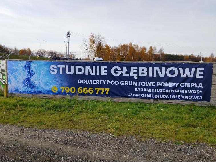 Baner reklamowy z napisem 'Studnie głębinowe' i numerem telefonu na betonowym ogrodzeniu, otoczony zielenią i drzewami w tle.