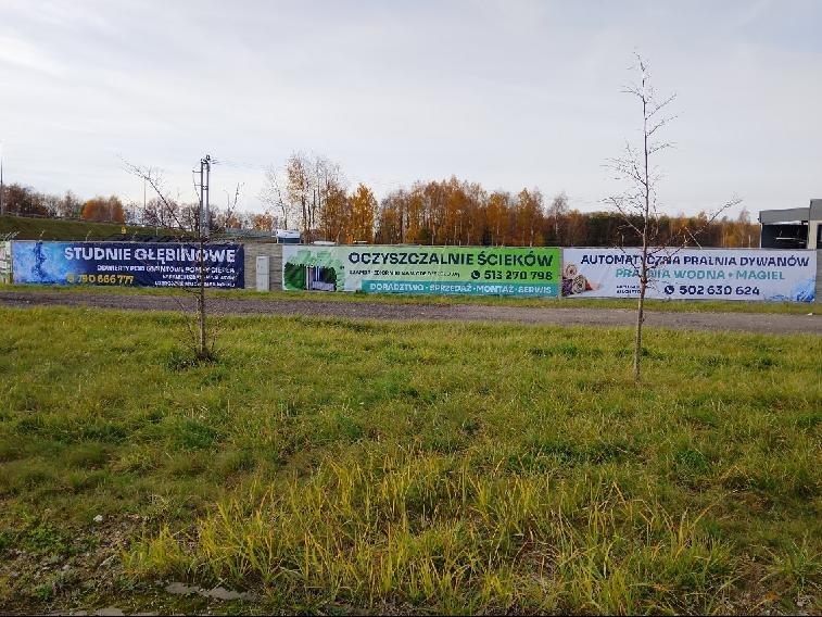 Banery reklamowe: studnie głębinowe, oczyszczalnie ścieków i pralnia dywanów. Widok na łąkę i drzewa w tle. Tablice z numerami telefonów.