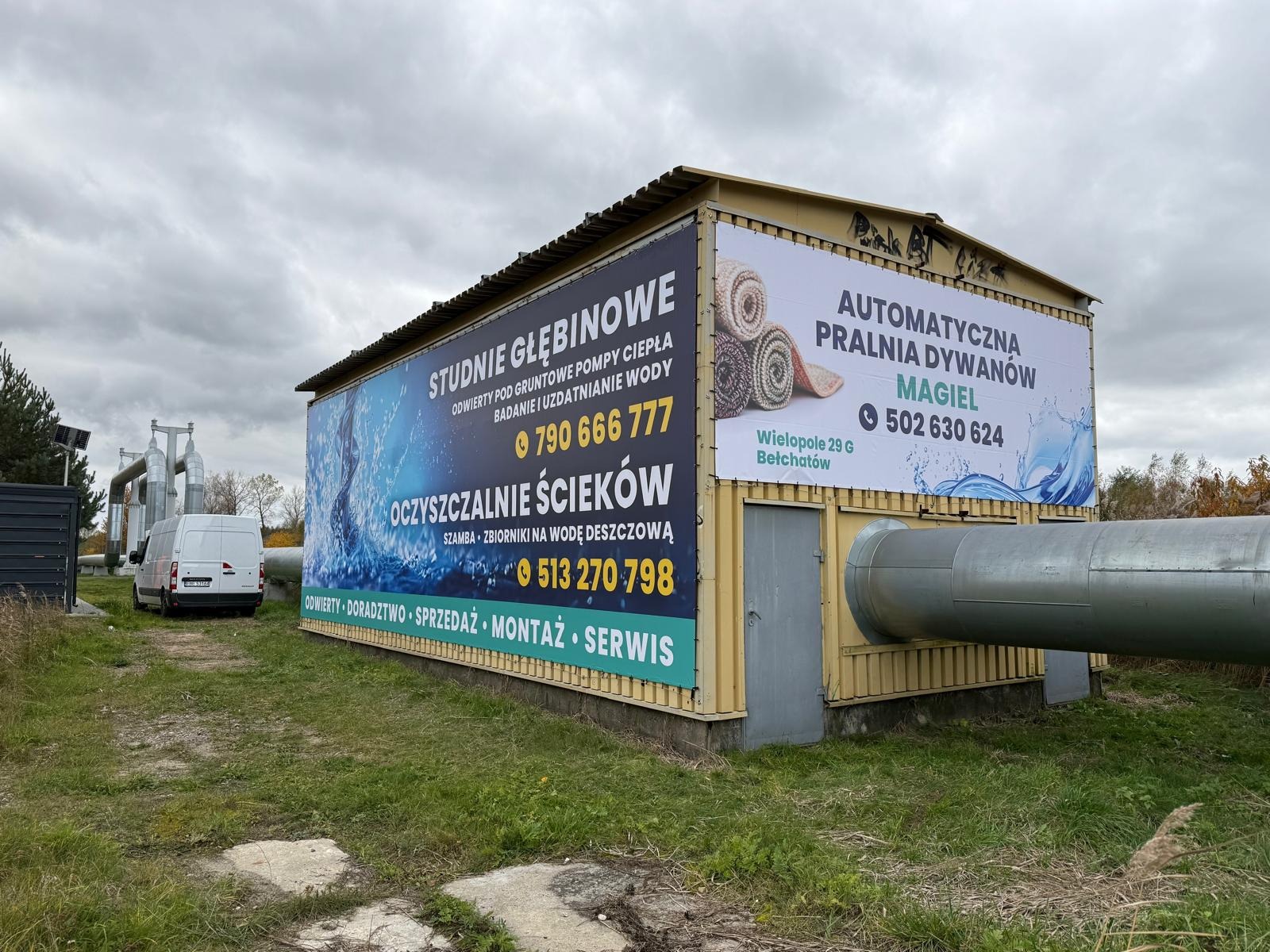 Budynek z reklamami studni głębinowych, oczyszczalni ścieków i pralni dywanów. Widoczny biały van i instalacje rurowe. Pochmurne niebo w tle.