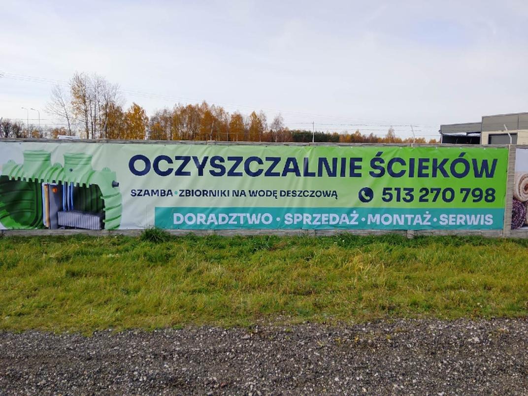 Baner reklamowy oczyszczalni ścieków, szamb i zbiorników na deszczówkę z numerem telefonu, widoczny na tle zieleni i drzew.