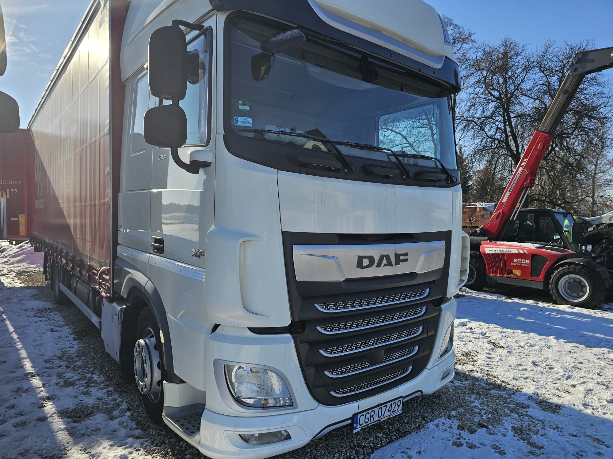 Biała ciężarówka DAF XF na ośnieżonym terenie, z czerwoną ładowarką teleskopową Massey Ferguson w tle. Widoczna tablica rejestracyjna i logo DAF.