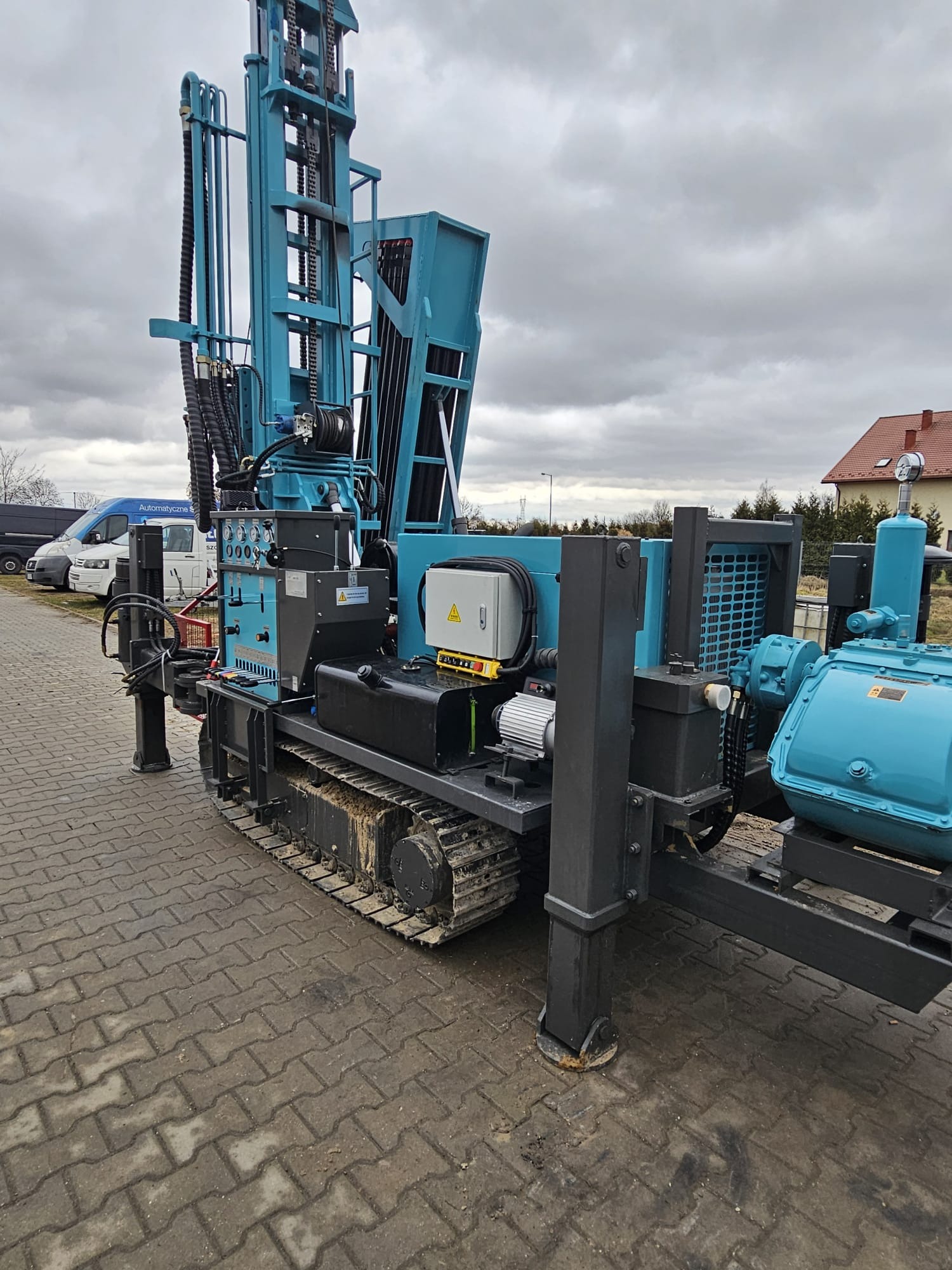 Mobilna wiertnica studni głębinowych na gąsienicach, w kolorze niebiesko-czarnym, zaparkowana na kostce brukowej. Widoczne elementy sterujące i przewody hydrauliczne.