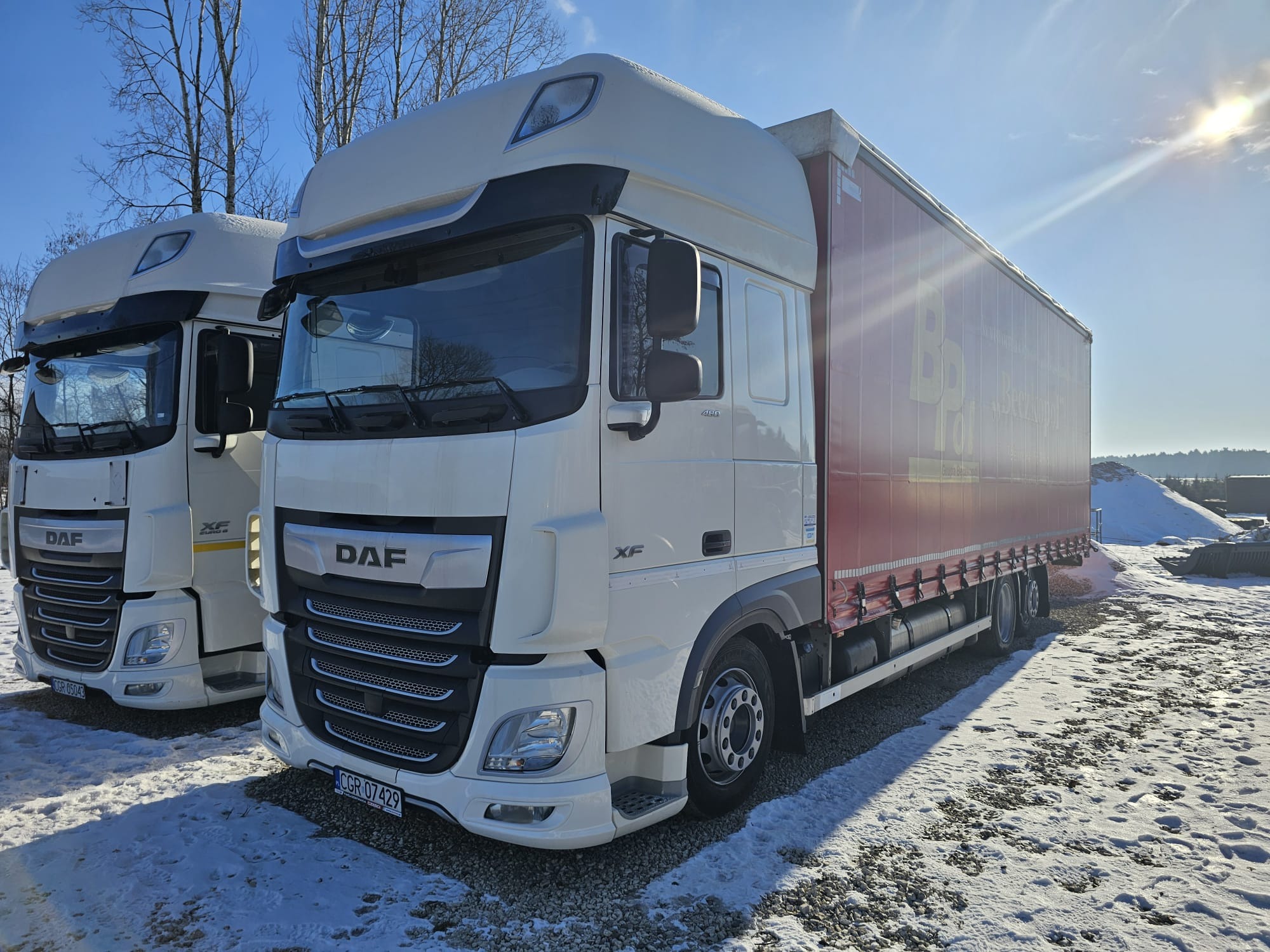 Biała ciężarówka DAF XF z czerwoną naczepą na zimowym placu, obok druga ciężarówka. Słońce w kadrze, widoczne tablice rejestracyjne. Transport drogowy.