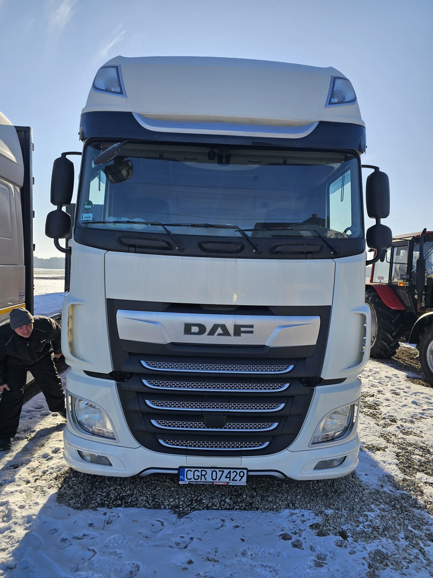 Biała ciężarówka DAF z przodu, z widoczną tablicą rejestracyjną i osobą sprawdzającą oponę, na tle zaśnieżonego terenu i częściowo widocznego traktora.
