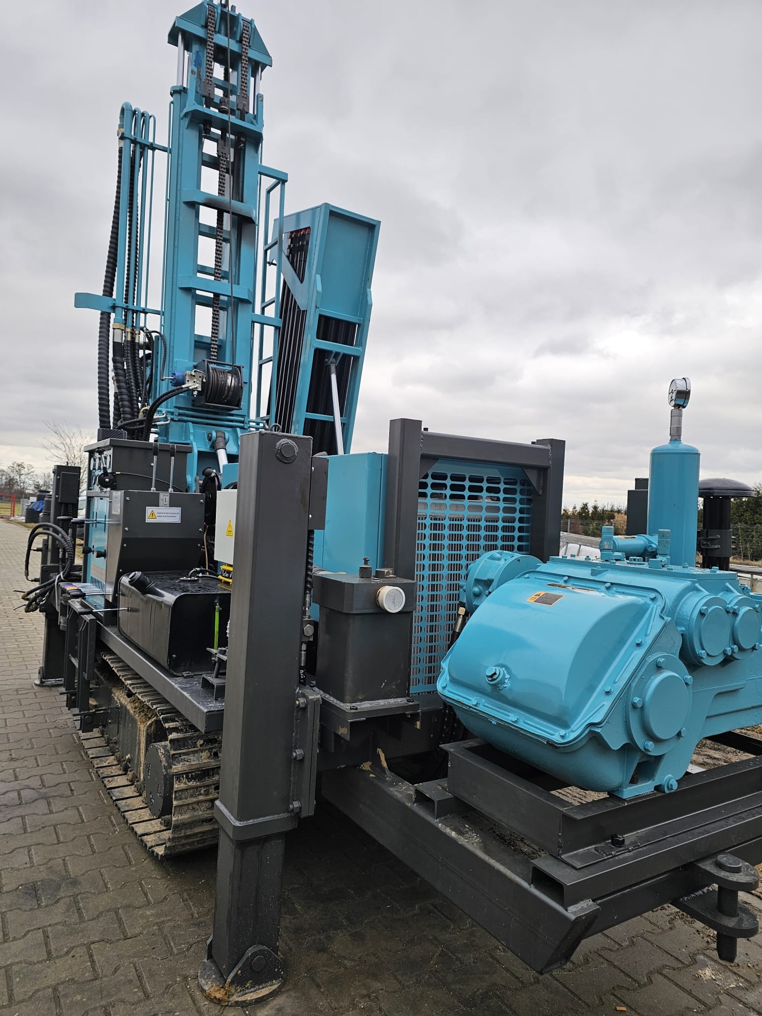 Niebiesko-czarna wiertnica na gąsienicach, widok z boku, pochmurne niebo w tle. Sprzęt do wiercenia studni głębinowych na kostce brukowej.