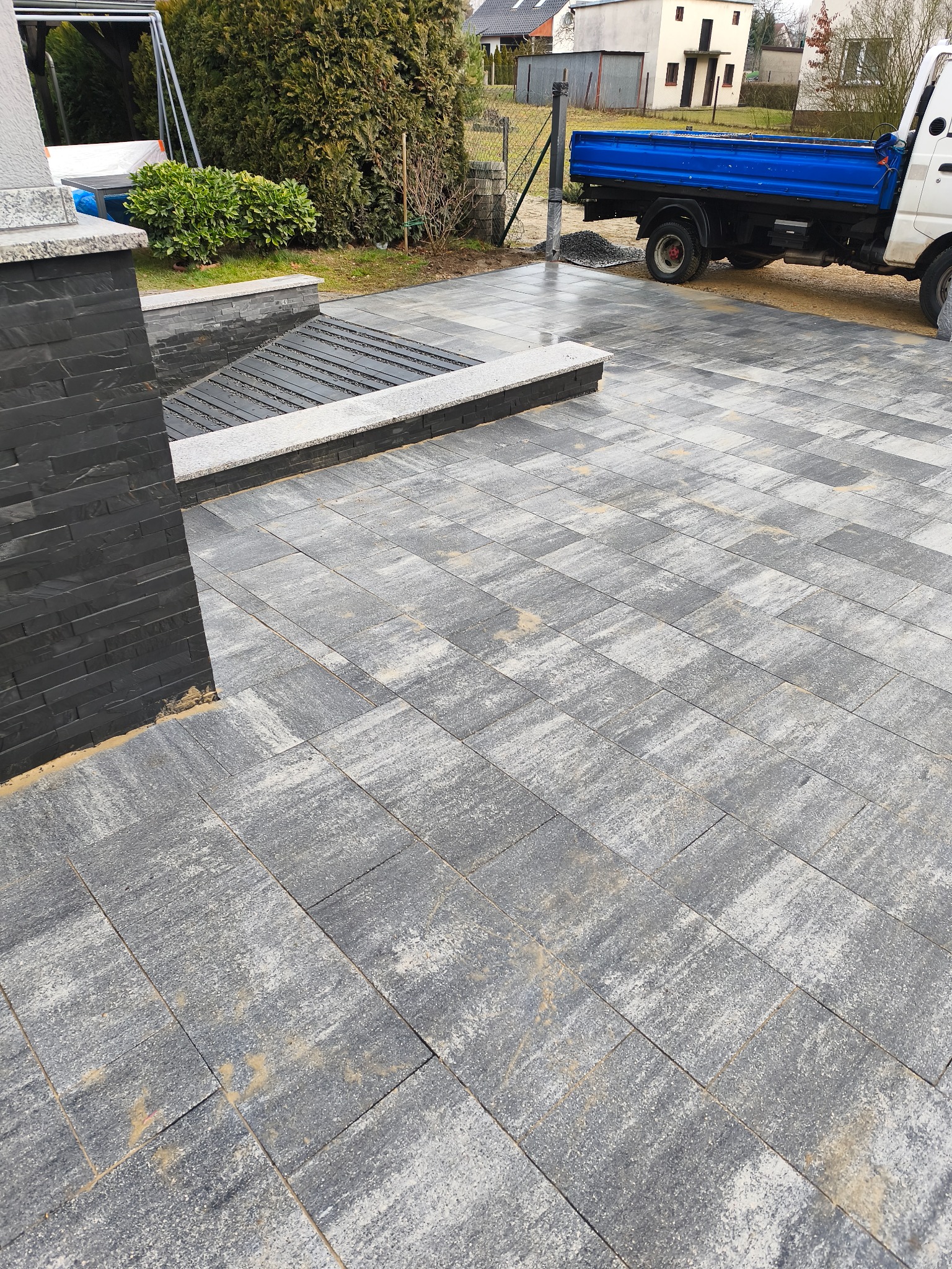 Ułożona szara kostka brukowa na posesji, z elementami dekoracyjnymi z ciemnego kamienia. W tle widoczny samochód dostawczy i zabudowa jednorodzinna.