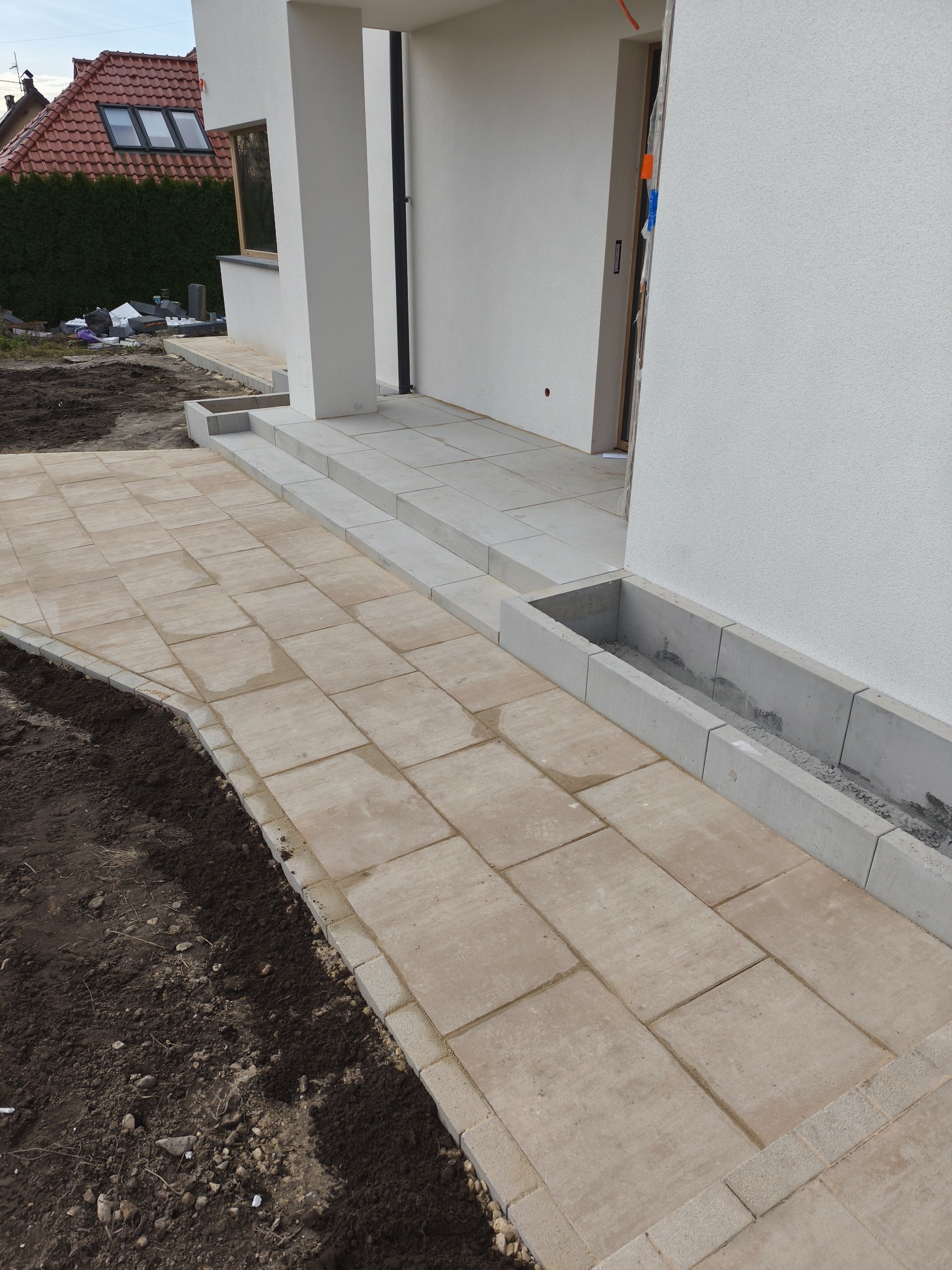 Jasne, prostokątne płyty chodnikowe prowadzą do wejścia domu z betonowymi schodami i korytkiem drenażowym. Obrzeża z małej kostki. Surowy grunt po lewej.