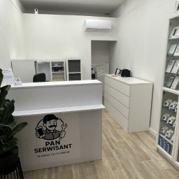KM Karol Misztal - Wnętrze punktu serwisowego telefonów: biała lada z logo 'Pan Serwisant', ekspozycja akcesoriów w gablotach, minimalistyczny wystrój w jasnych barwach.