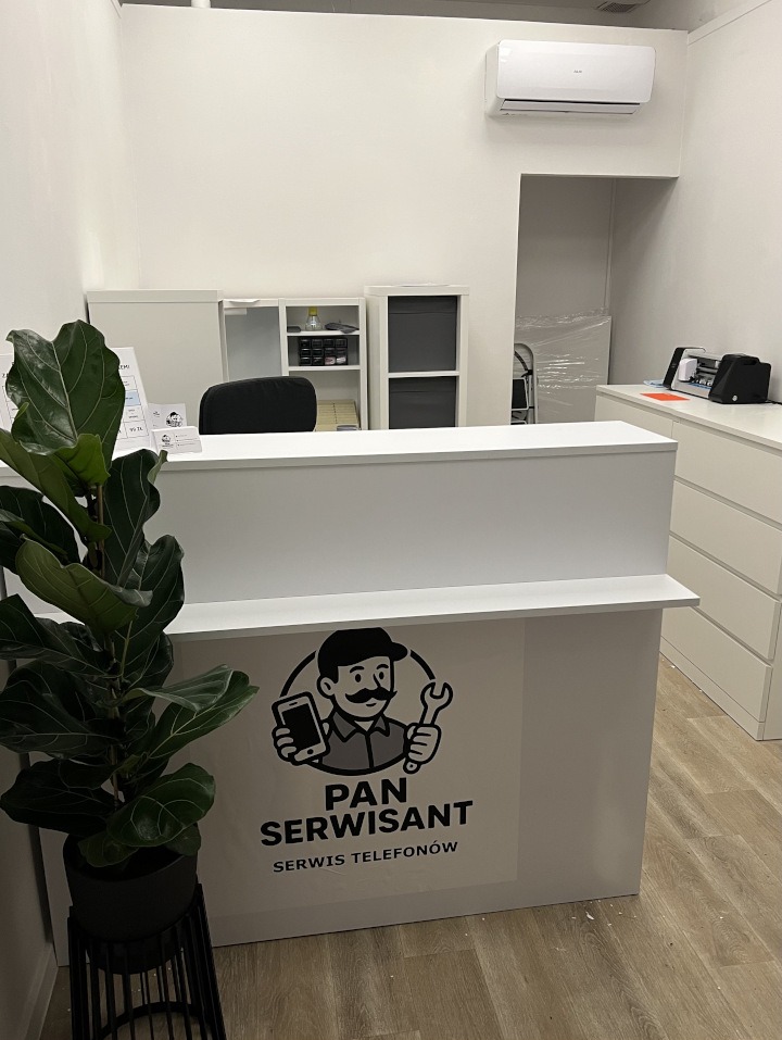 Recepcja serwisu telefonów 'Pan Serwisant' w Krakowie. Na ladzie logo firmy z wizerunkiem serwisanta. W tle białe meble i klimatyzator. Minimalistyczny wystrój.