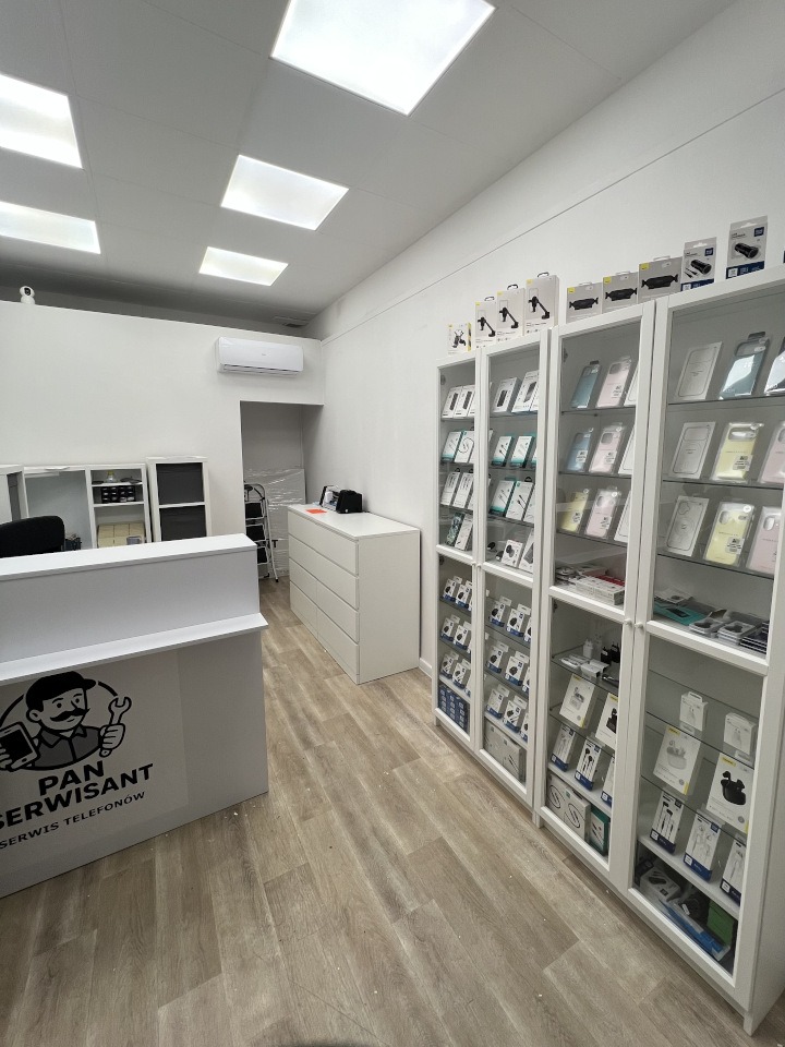 Wnętrze punktu serwisowego telefonów: białe meble, gabloty z akcesoriami, lada z logo 'Pan Serwisant'. Jasne oświetlenie, minimalistyczny design.