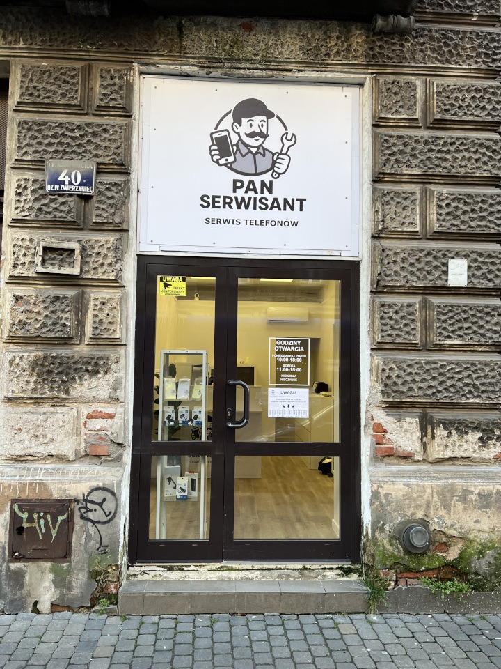 Wejście do serwisu telefonów 'Pan Serwisant' z logo przedstawiającym postać z telefonem i kluczem. Widoczny adres i godziny otwarcia na drzwiach.