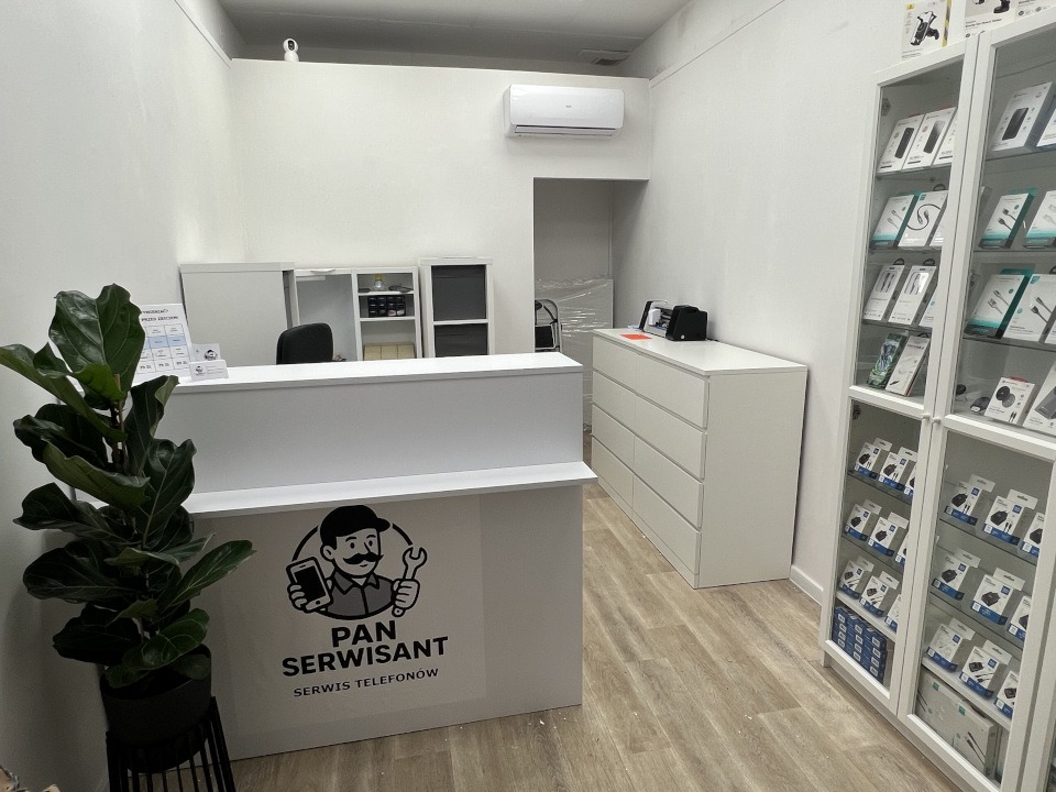 Wnętrze punktu serwisowego telefonów: biała lada z logo 'Pan Serwisant', ekspozycja akcesoriów w gablotach, minimalistyczny wystrój w jasnych barwach.
