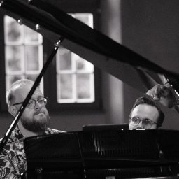 Vitalii Ishchenko - Czarno-białe zdjęcie dwóch mężczyzn w okularach grających na fortepianie. Jeden w kwiecistej koszuli, drugi zza instrumentu. Scena z koncertu lub sesji nagraniowej.