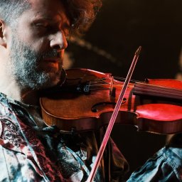 Vitalii Ishchenko - Mężczyzna z brodą gra na skrzypcach w skupieniu, oświetlony ciepłym światłem. W tle rozmyte światła i sylwetka postaci. Ujęcie z bliska, podkreślające emocje muzyka.